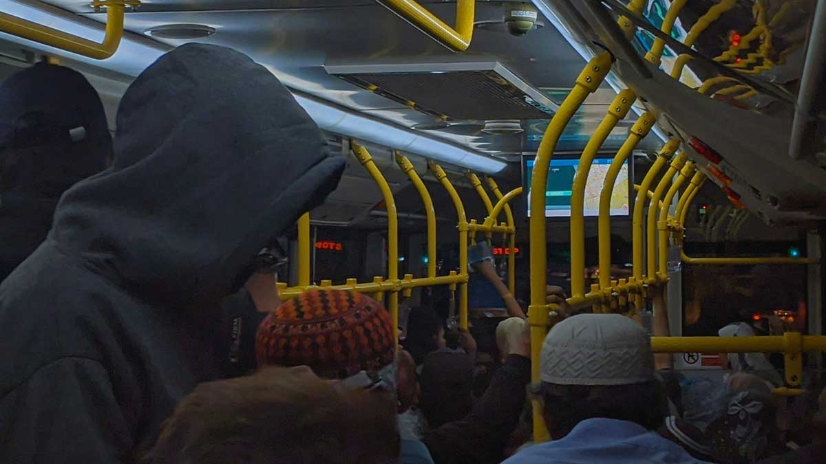 Eine überfüllte Busfahrt bei Nacht. Viele Menschen stehen dicht gedrängt im Gang, einige halten sich an den gelben Haltestangen fest. Eine Person trägt einen dunklen Kapuzenpulli, andere haben Mützen oder Kopfbedeckungen auf. Im Hintergrund ist ein Bildschirm mit einer Karte zu sehen. Viele Reisende wünschen sich einen Führerschein statt einer Fahrkarte