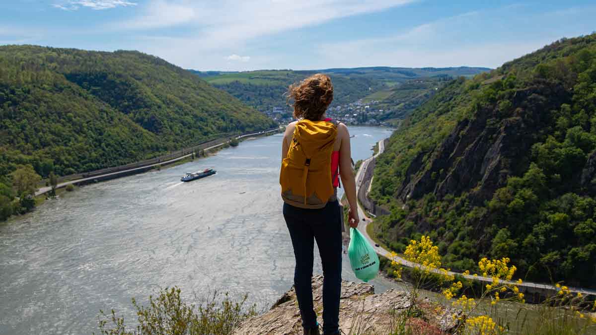 Wanderin mit Rucksack blickt vom Loreley-Felsen auf den Rhein, in der Hand eine grüne Mülltüte der Aktion ‚Mach mit – Nimm mit‘