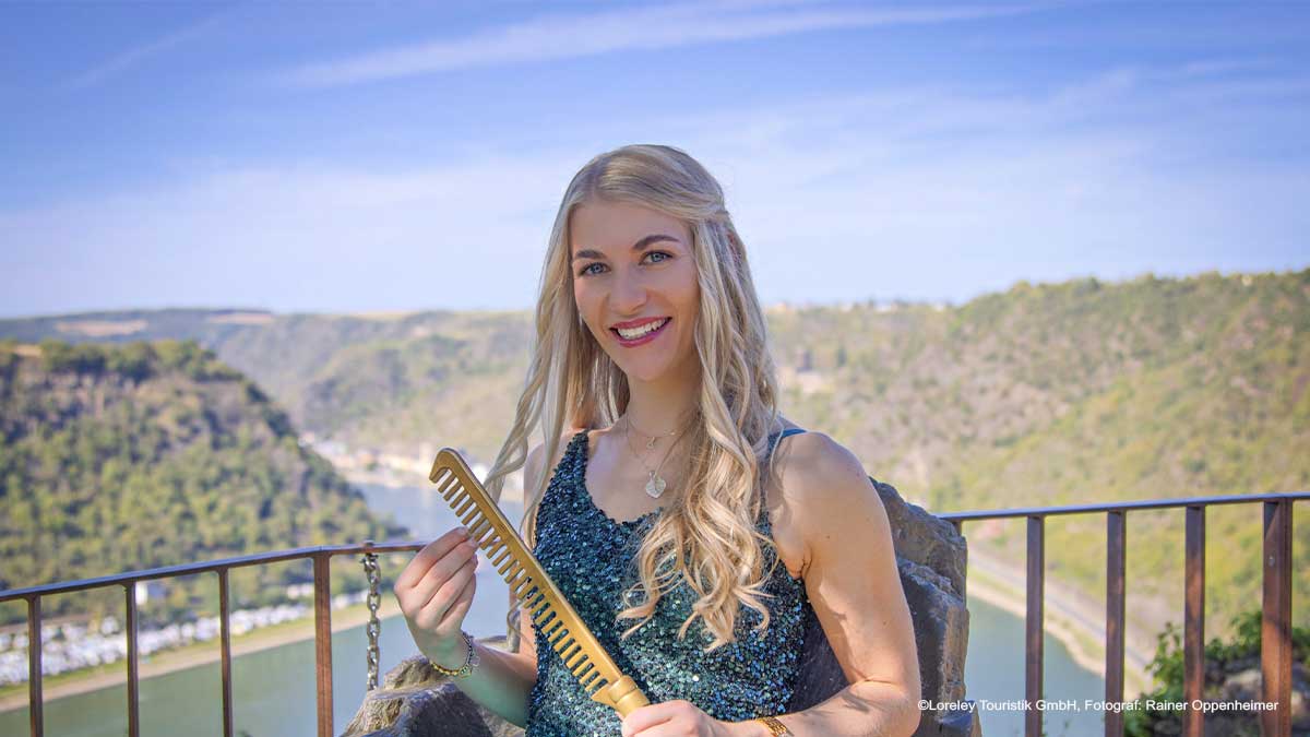 Eine junge Frau mit langen, blonden Haaren lächelt in die Kamera. Sie trägt ein glitzerndes, dunkelgrünes Kleid und hält einen goldfarbenen Kamm in der Hand. Im Hintergrund fließt der Rhein durch das Mittelrheintal, umgeben von bewaldeten Hügeln.