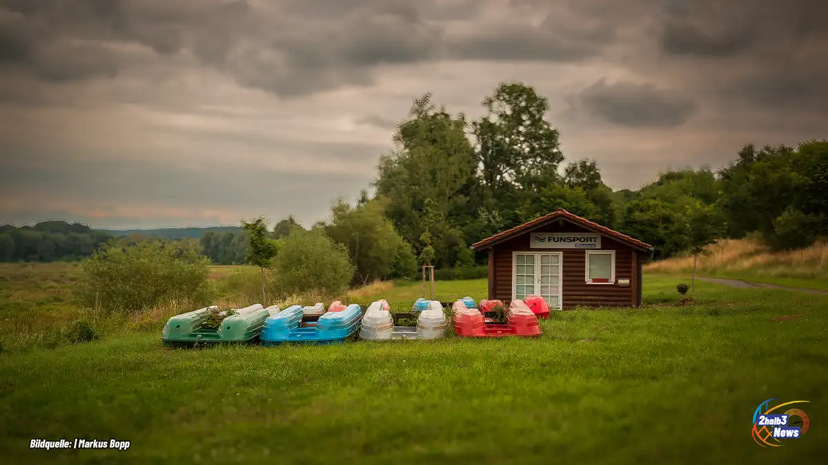 Trockengelegter Wiesensee: Bunte Tretboote stehen ungenutzt auf einer Wiese vor einer kleinen Verleihhütte, im Hintergrund bewaldete Hügel unter grauem Himmel.