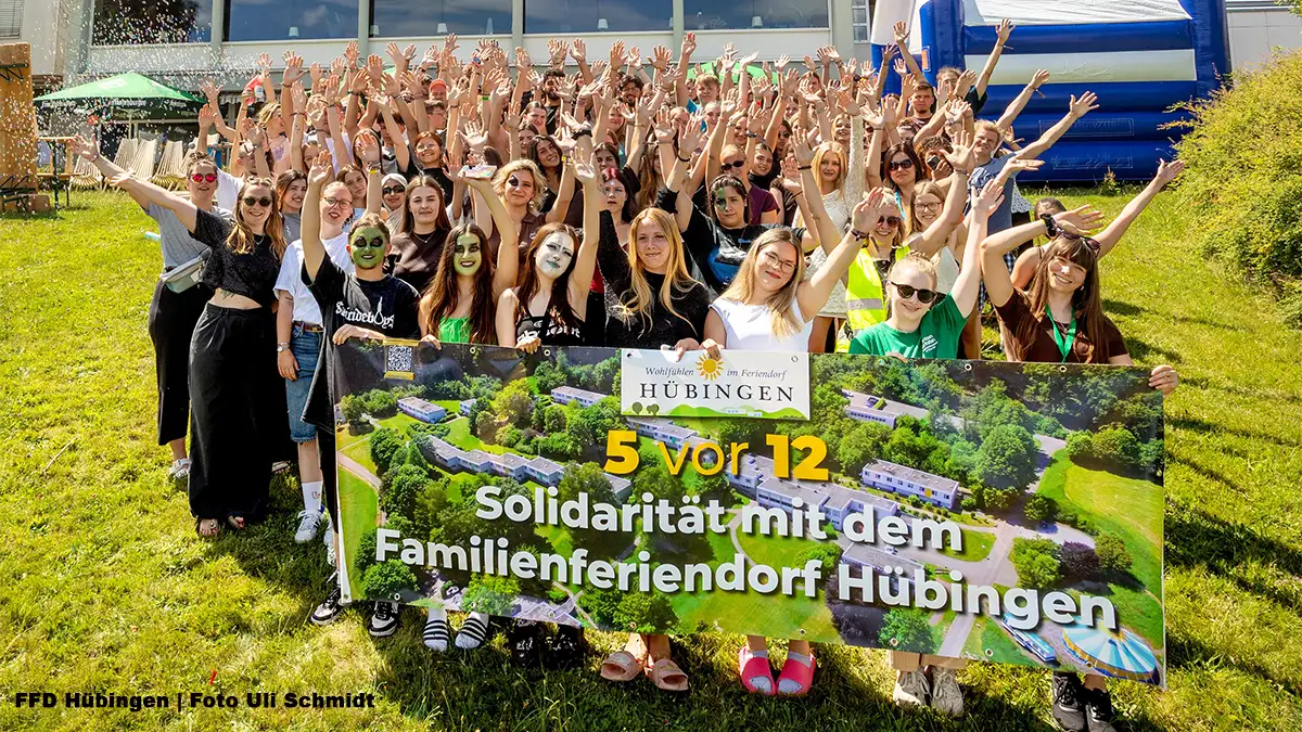 Zahlreiche junge Freiwilligendienstleistende stehen mit erhobenen Händen vor dem Familienferiendorf Hübingen und halten ein Banner mit der Aufschrift „5 vor 12 – Solidarität mit dem Familienferiendorf Hübingen“.